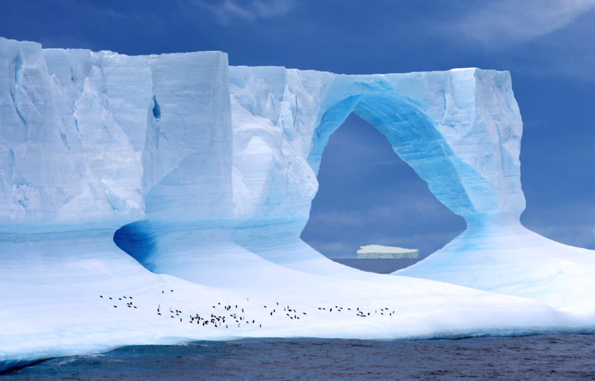 Ein bogenförmiger Eisberg vor dem eine Kolonie Kaiserpinguine steht.